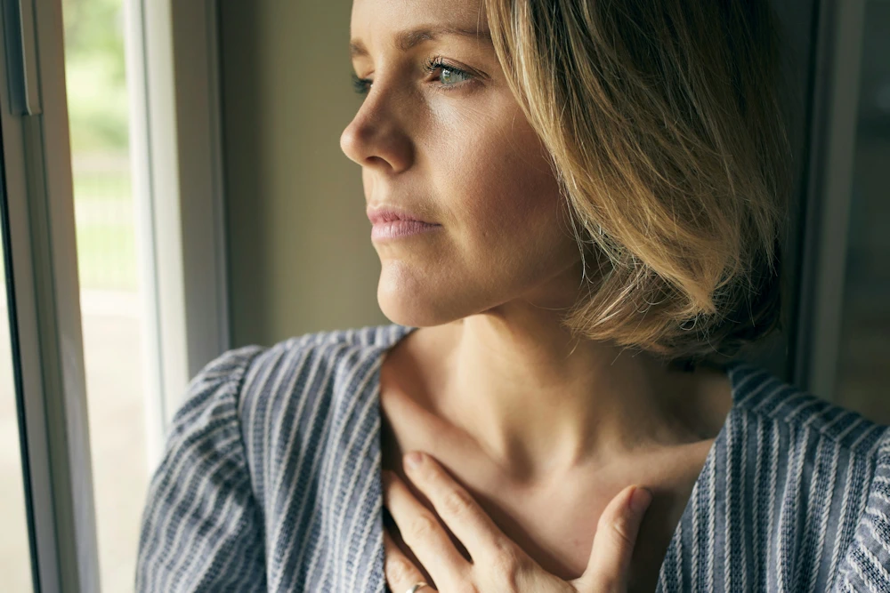 Vrouw kijkt in gedachten uit het raam met hand op haar borst, symbool voor innerlijke veiligheid
